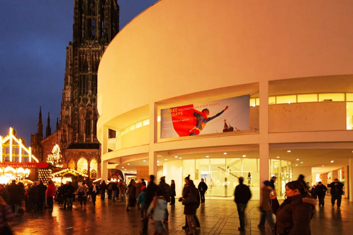 Crowd of pedestrians at night in a plaza beside a curved white modern building; a Gothic cathedral tower looms in the background with festive lights.