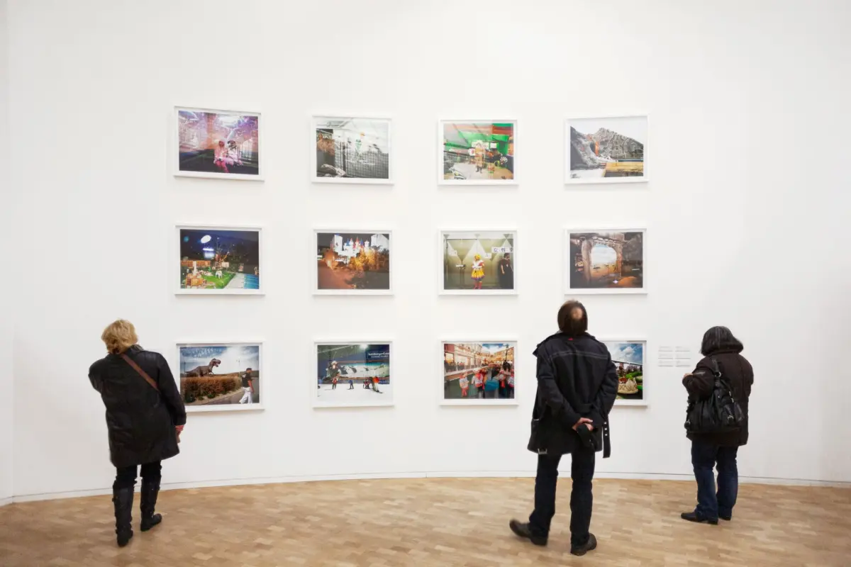 Three visitors stand facing a wall of eight framed photographs arranged in two rows of four in an art gallery.