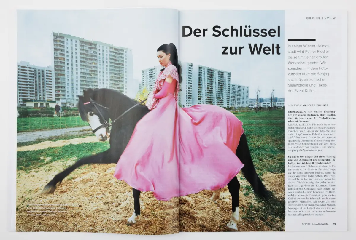Person riding a black horse in a pink dress on a dirt field with apartment buildings in the background; magazine spread header Der Schlüssel zur Welt.