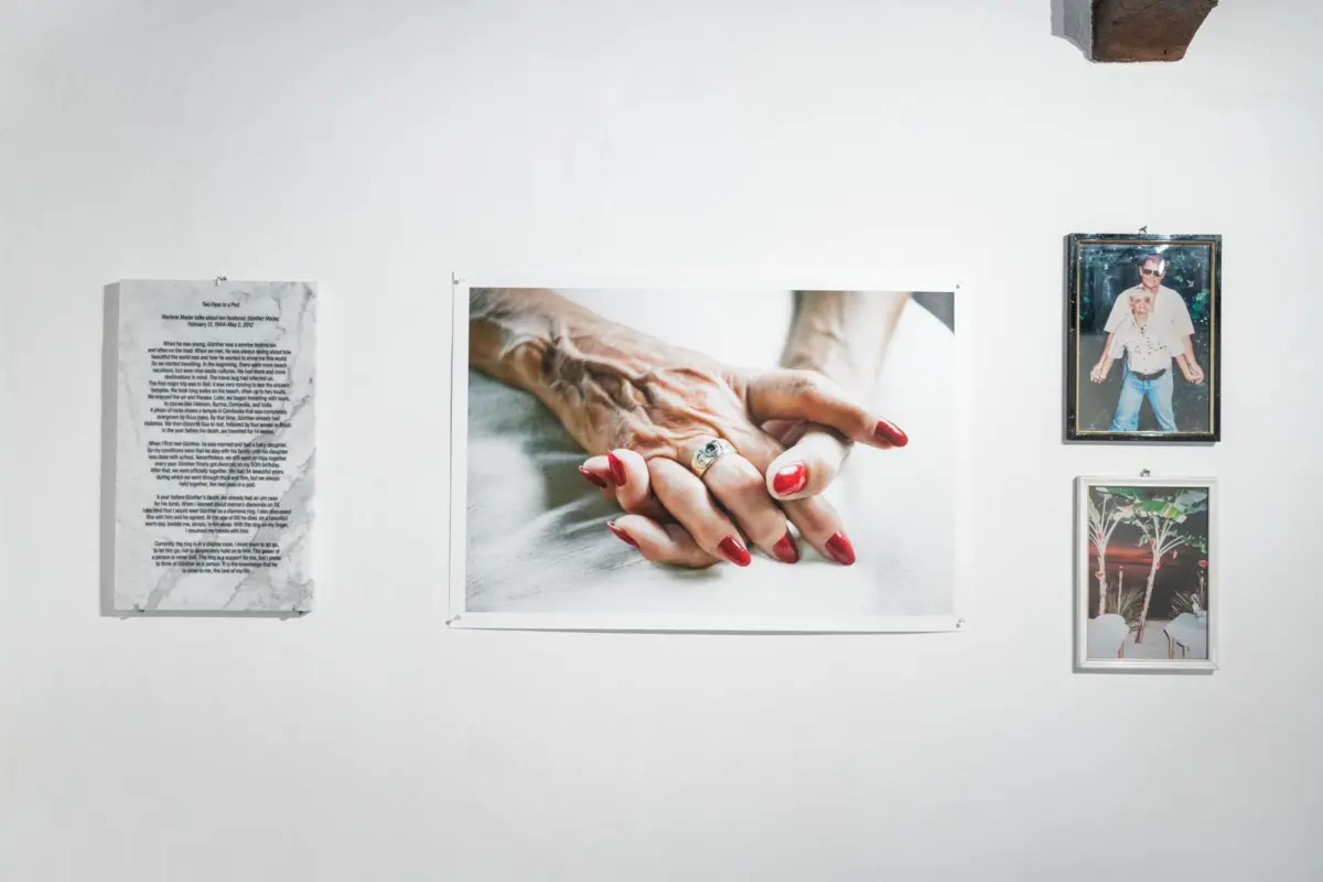 Interlaced hands with red nails in a large photograph on a white gallery wall.