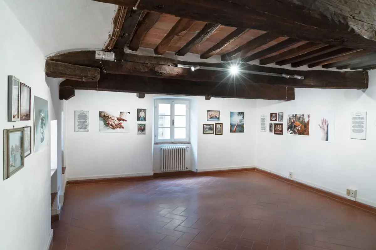 Interior of a small gallery with exposed dark wooden beams, brick arch, track lighting, and framed photographs on white walls; window and radiator at the back.