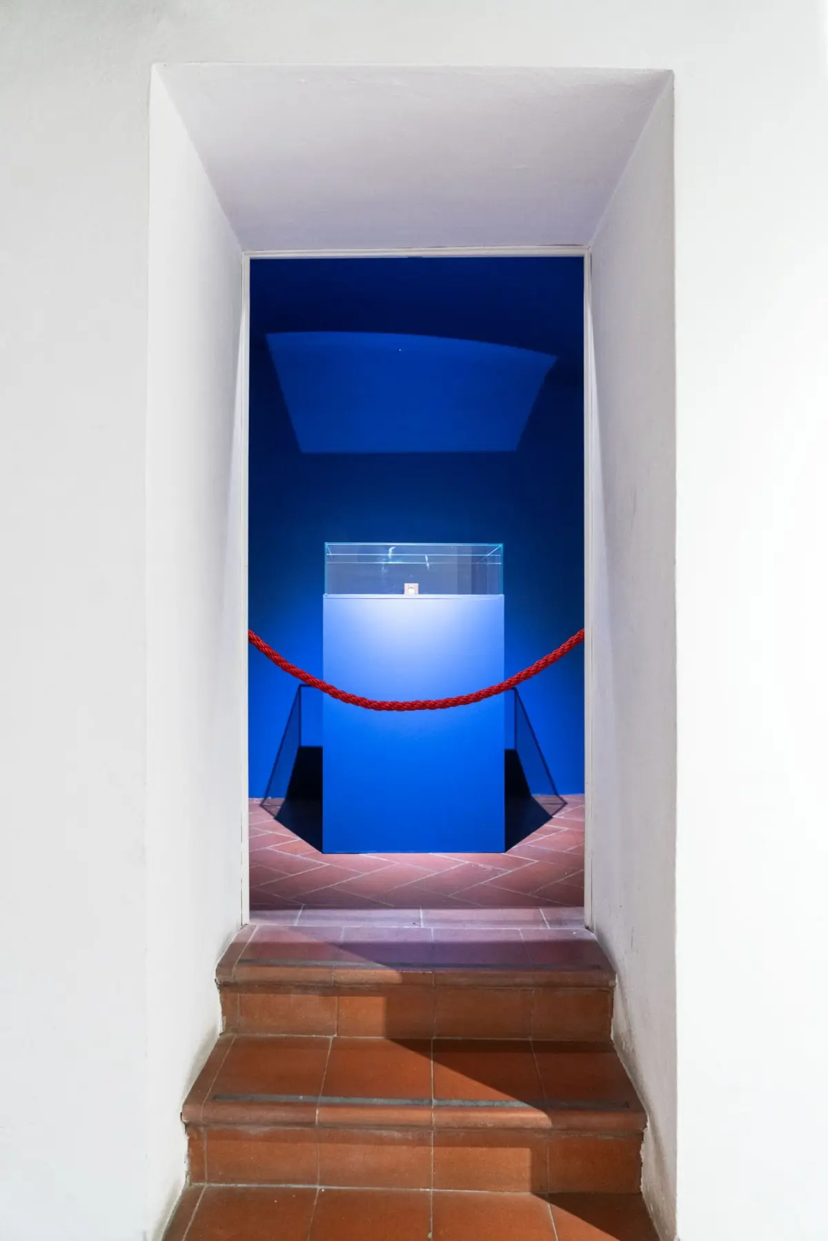 Through white doorway, blue-lit room displays glass display case atop blue pedestal, roped off with red barrier.