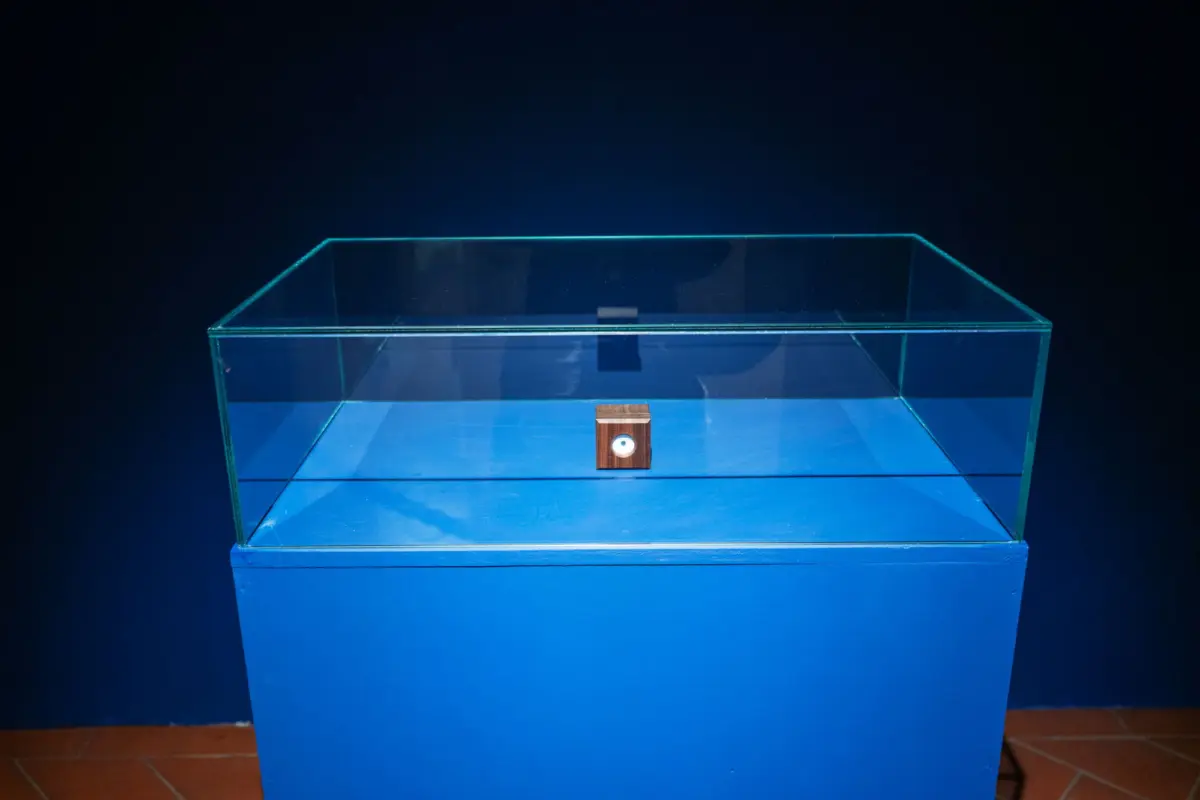 Glass display case with cyan edges on a blue pedestal against a dark background, housing a small wooden cube with a white circular inset.