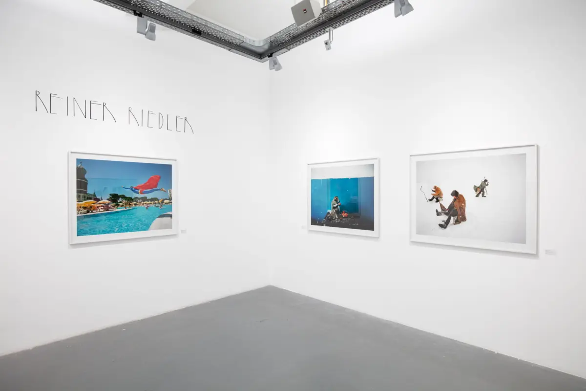 Gallery corner displaying three framed photographs on white walls; overhead metal track lighting; wall text reads REINER NEDLER.