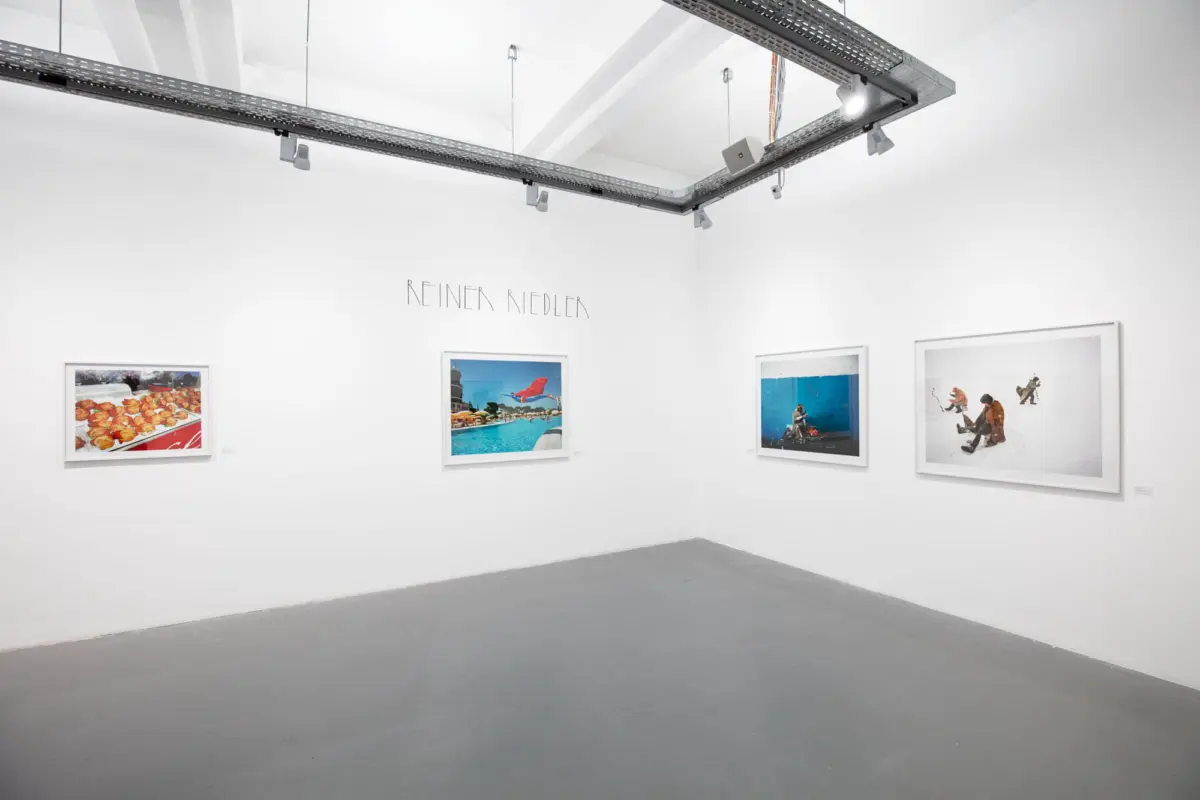 White-walled gallery with four framed photographs, prominent 'REINER RIEDLER' text on the wall, and exposed overhead track lighting.