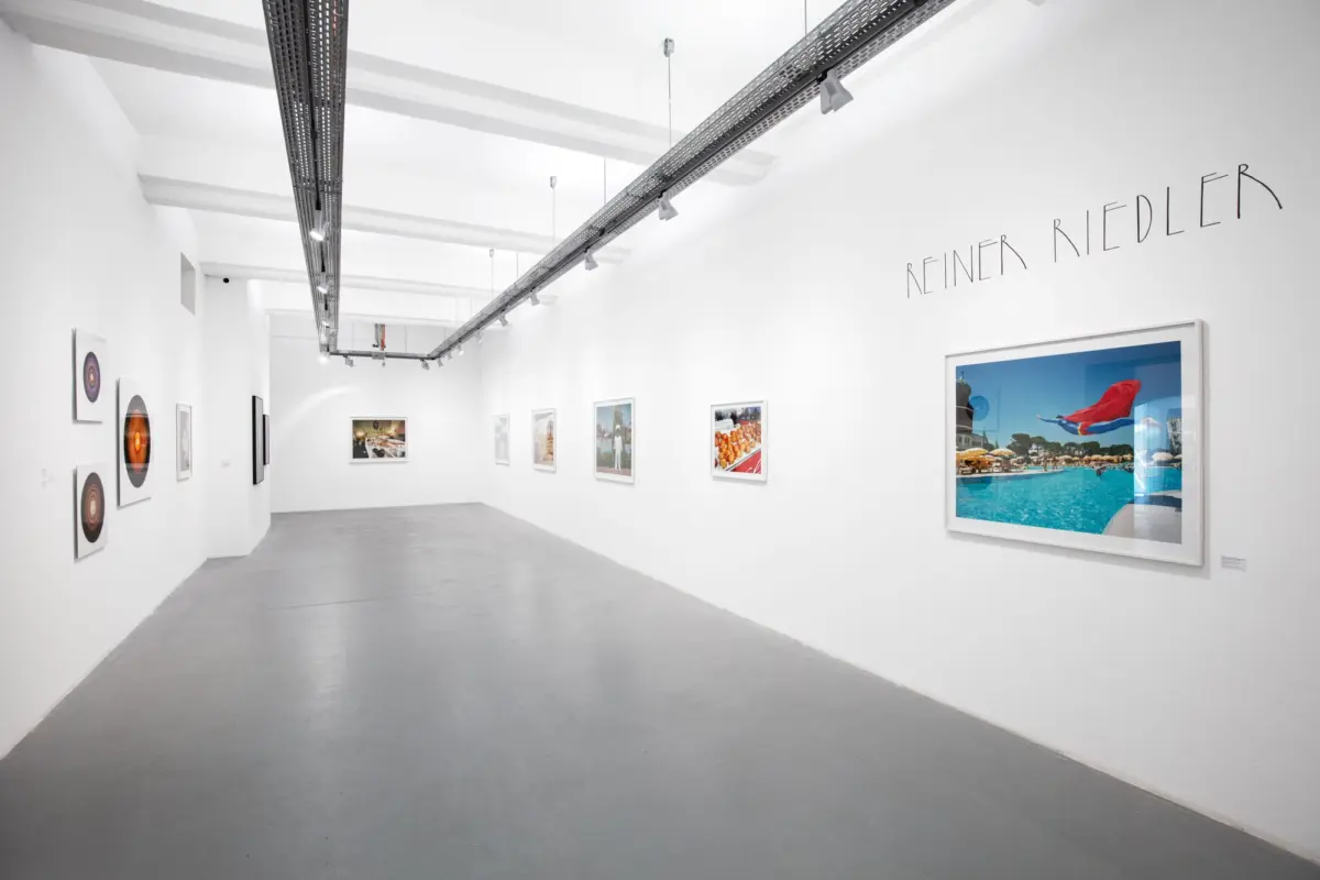 White-walled art gallery with ceiling track lighting; framed photographs along the walls; artist name 'REINER RIEDLER' visible on the right.