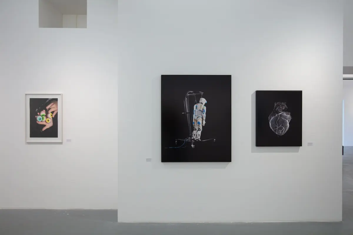 White-walled gallery with artworks: left hands holding colorful eyeballs; center astronaut on a wheeled platform; right white-line heart on black.