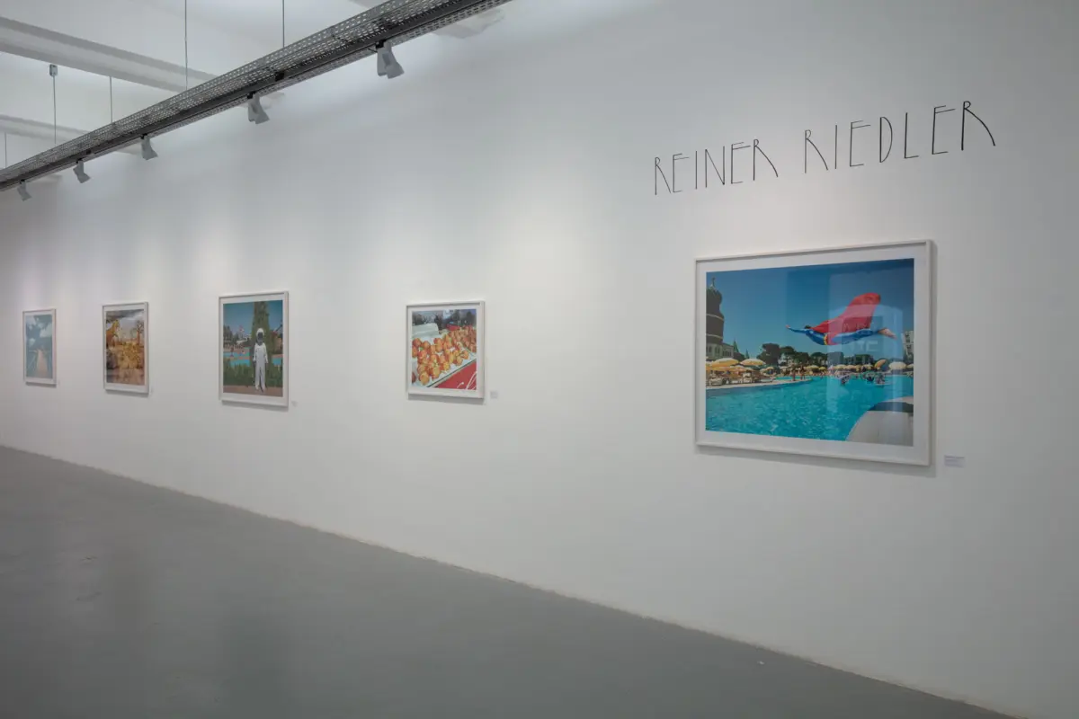White-walled gallery with six framed photographs along the wall, overhead track lighting, and the name 'REINER RIEDLER' displayed prominently.