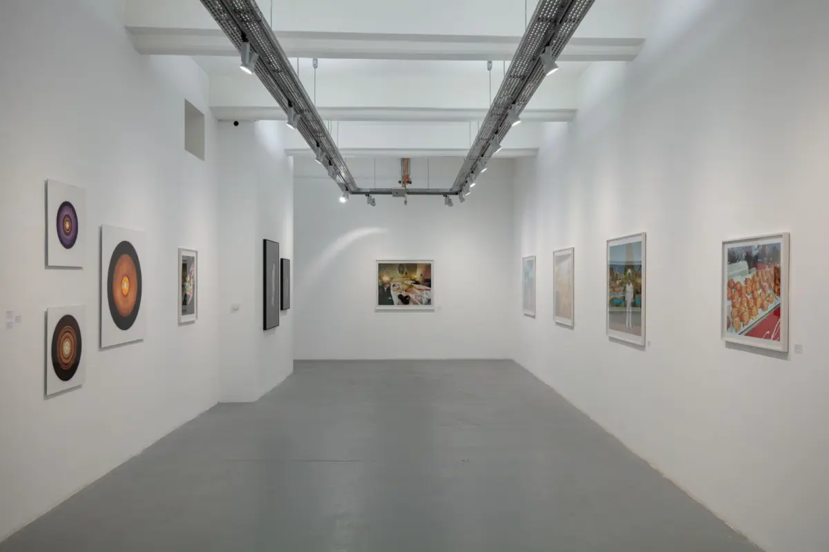 White-walled art gallery corridor; left wall displays circular abstract prints, right wall shows framed photos under track lighting.