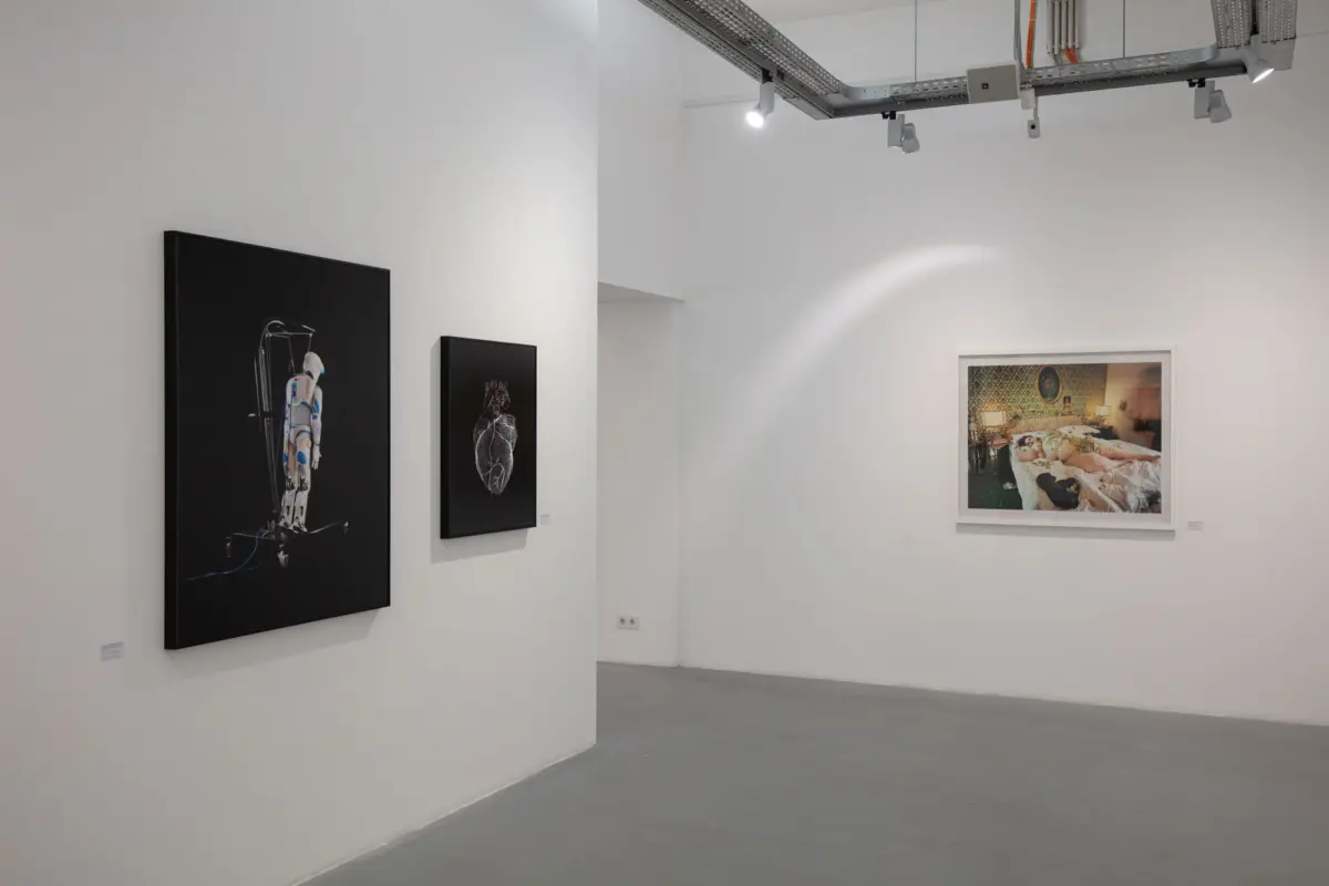Three framed photographs on white gallery walls: spacesuit figure on a stand, leaf close-up, and a bedroom scene.