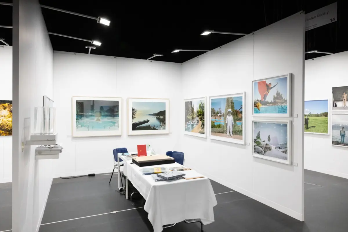 Art gallery booth with white walls, landscape photographs in frames, a table with documents, blue chairs, and a "Photon Gallery" sign.