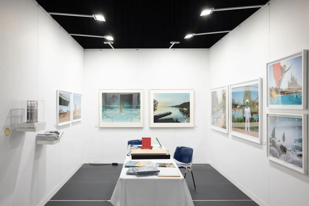 White-walled art gallery displaying framed travel photographs; central table with documents and two blue chairs under overhead lighting.