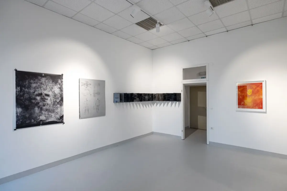 Art gallery interior with white walls; large black-and-white abstract left and a row of dark mini-works, plus an orange-framed piece on the right.