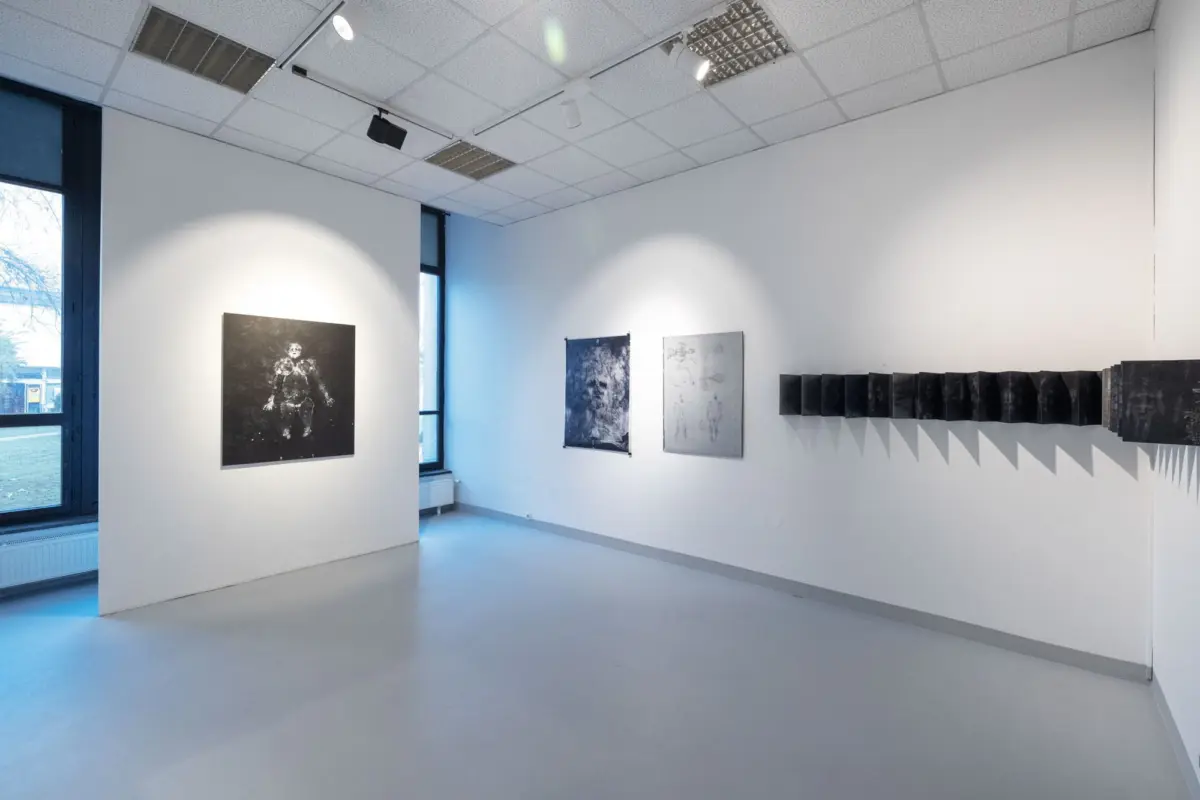 Art gallery interior featuring white walls, a large black-and-white portrait on the left, and a line of dark rectangular panels along the right wall.