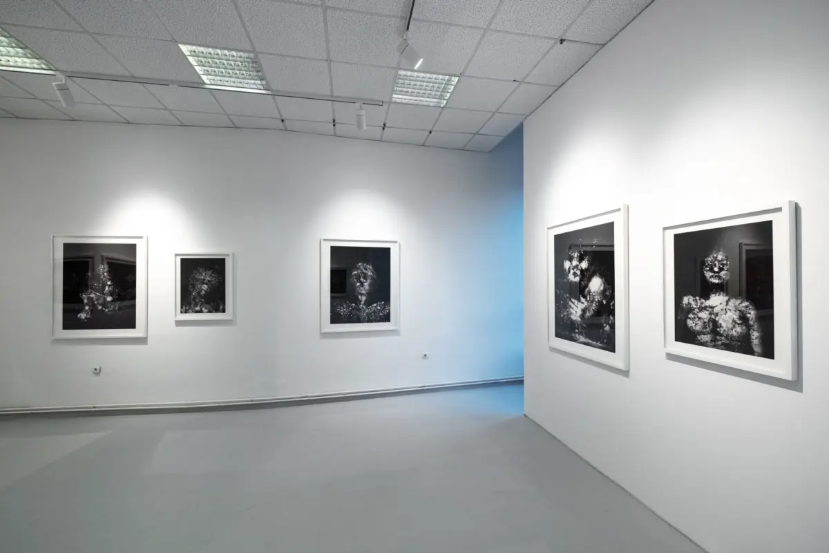 Art gallery interior with framed black-and-white photographs on white walls, gray floor, and overhead lights.