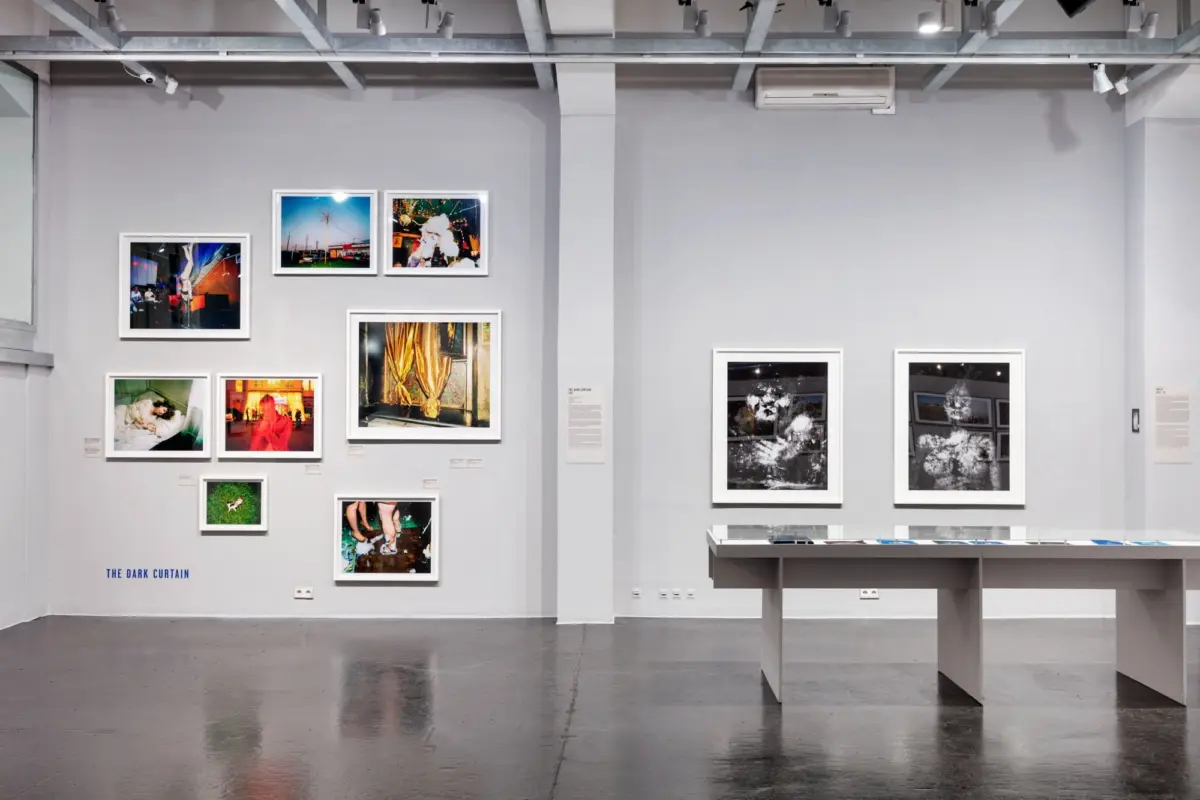 Interior of a contemporary art gallery with gray walls, framed photographs arranged in a grid, a white display table, and ceiling track lighting.