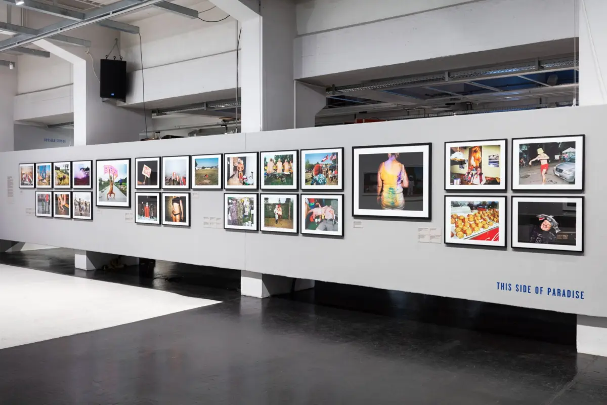 Indoor art gallery displaying a long white wall with numerous framed photographs in a grid, industrial ceiling above.