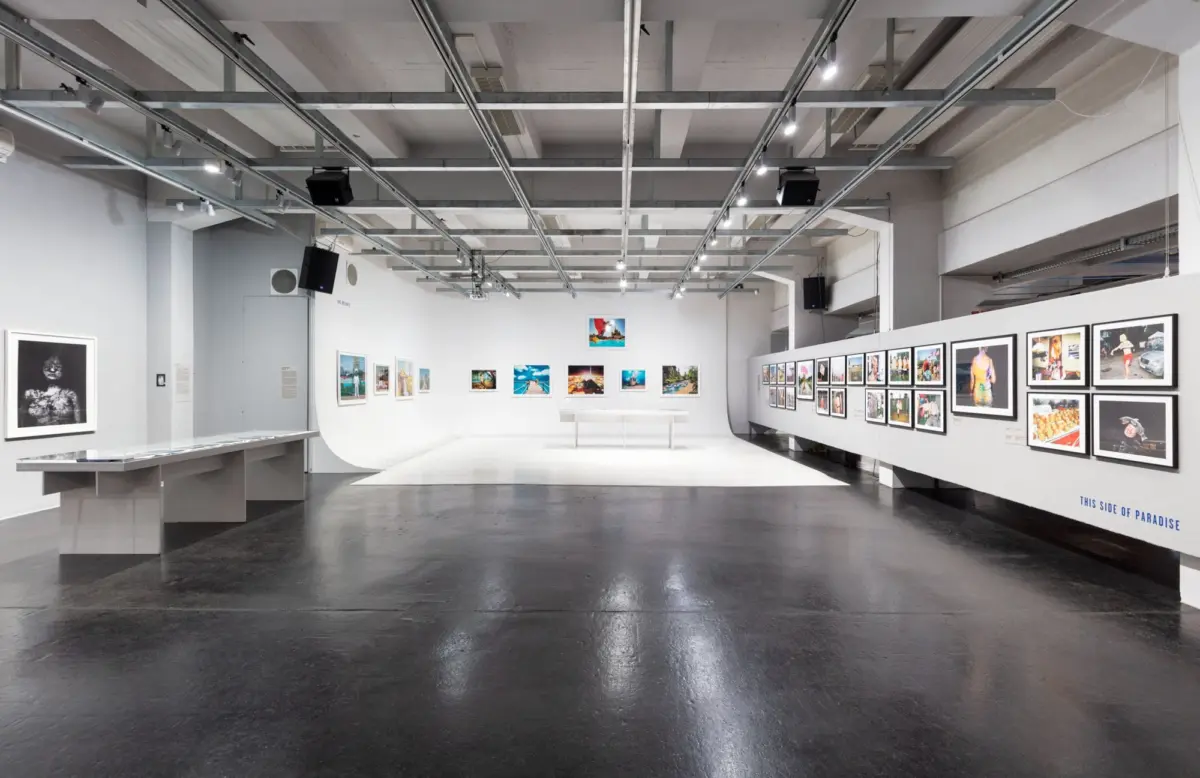 Spacious contemporary art gallery interior with rows of framed photographs on white walls, a dark polished floor, and exposed metal ceiling beams.