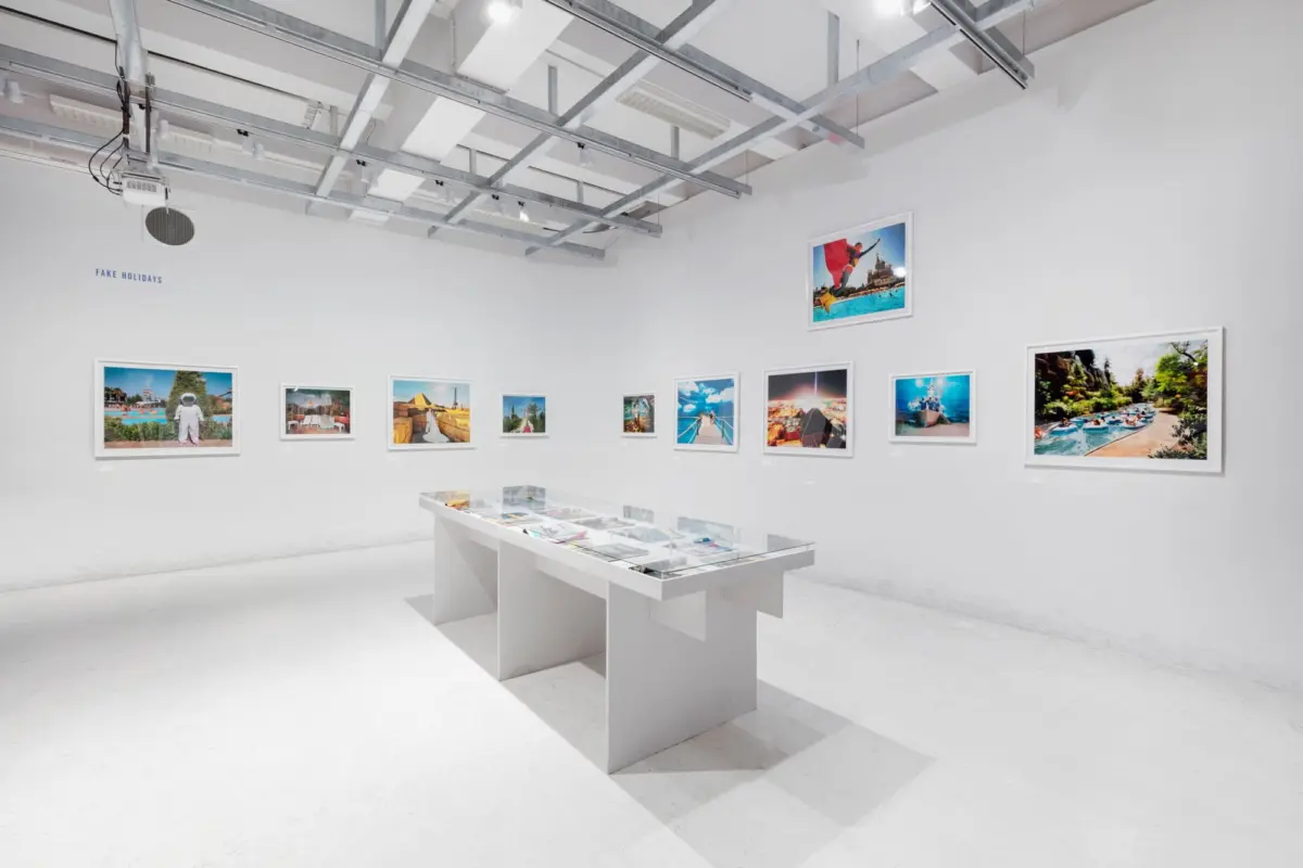White-walled gallery with framed travel photos along the walls, glass display tables, and an exposed metal ceiling grid; 'FAKE HOLIDAYS' on wall.