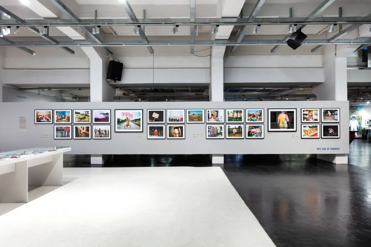 Industrial art gallery interior with a long row of framed photographs on a white wall, exposed metal beams, and track lighting overhead.
