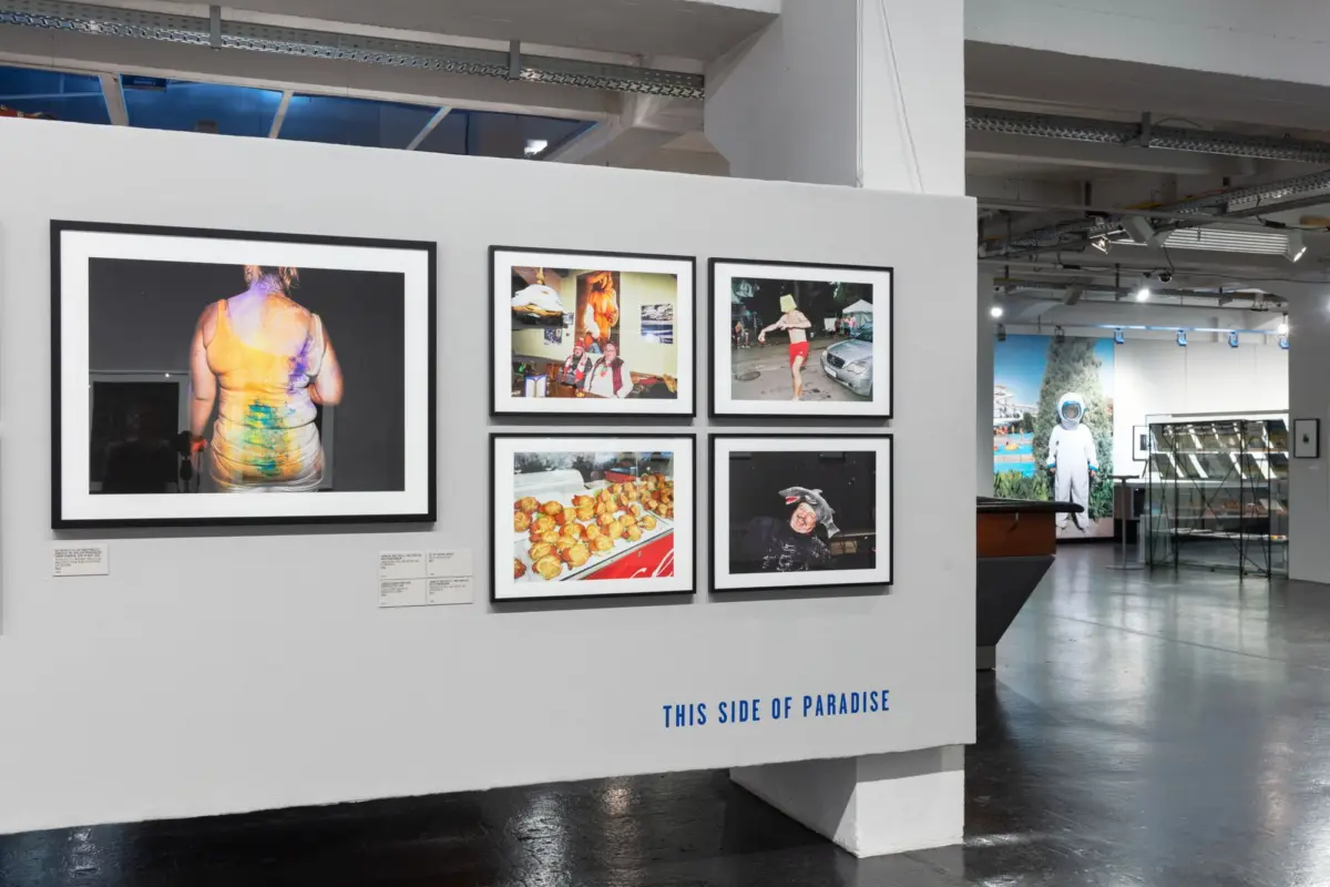 Art gallery wall with six framed photographs; back view of a paint-splattered person; blue text 'THIS SIDE OF PARADISE' visible.