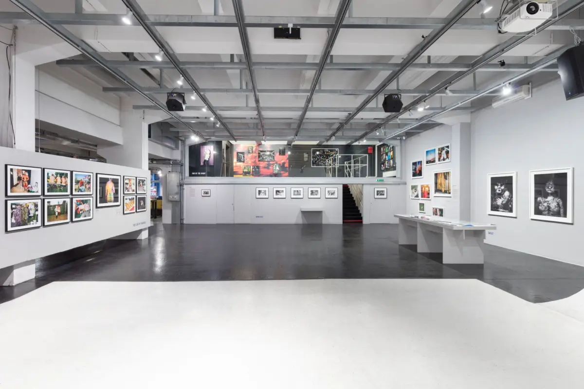 Interior of an art gallery with framed photographs along white walls, display tables, and a mezzanine staircase.
