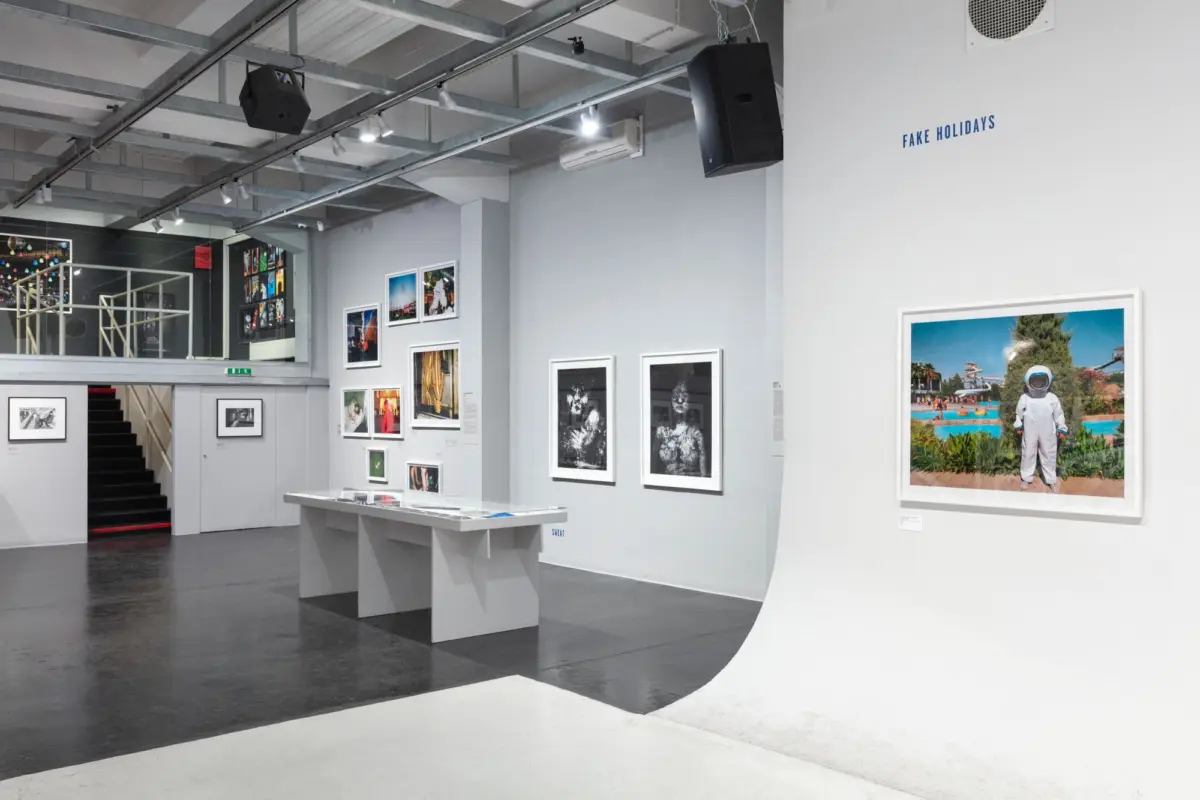 Art gallery interior with framed photographs on gray walls, exposed ceiling beams, curved white divider, and 'FAKE HOLIDAYS' sign.