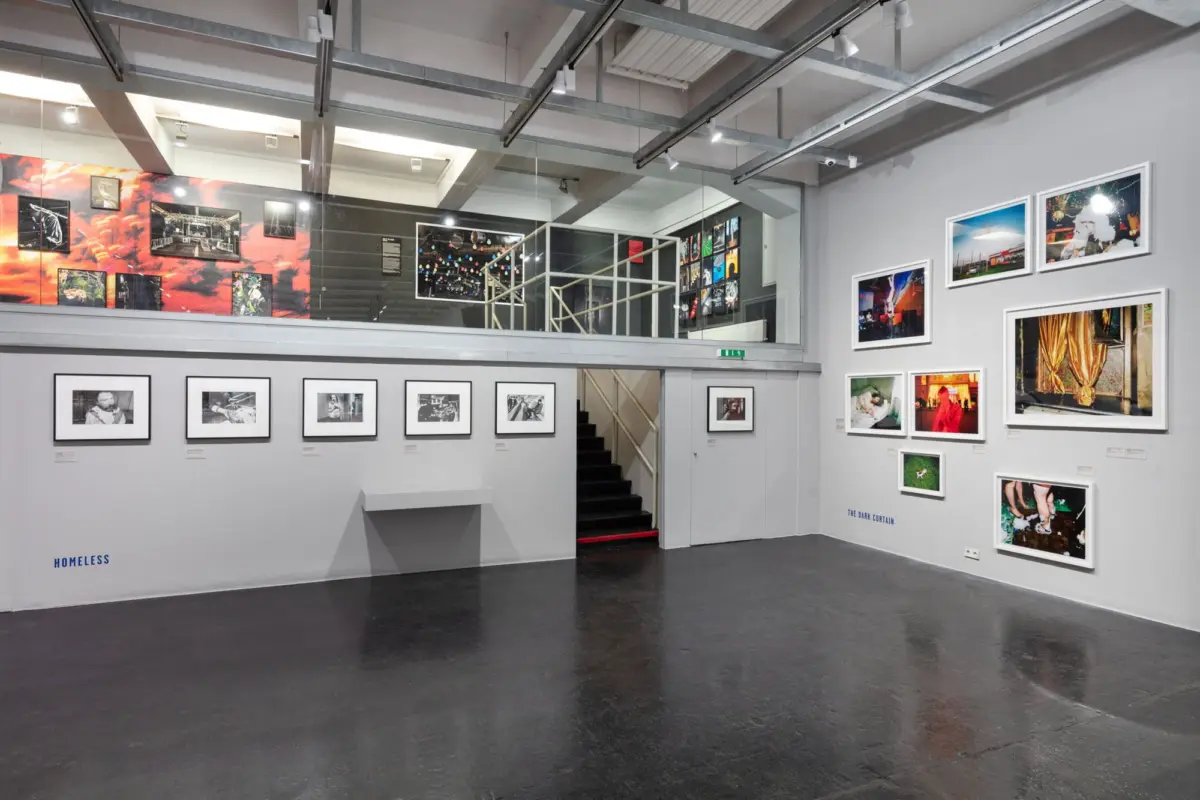 Interior of a contemporary art gallery: grey walls, numerous framed photographs, and a staircase leading to a mezzanine.