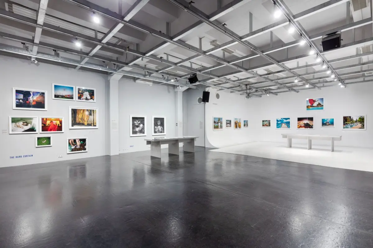 Contemporary art gallery interior with wall-mounted photographs, white walls, track lighting, and a central white display table.