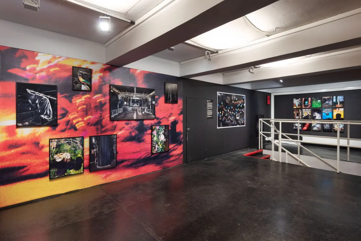 Art gallery interior featuring a bold red-orange mural wall with multiple framed photographs; metal railing along a stairwell.
