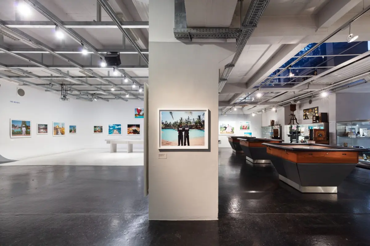 Museum gallery with framed travel photos on white walls and vintage boats on display; central pillar shows a tropical beach scene.
