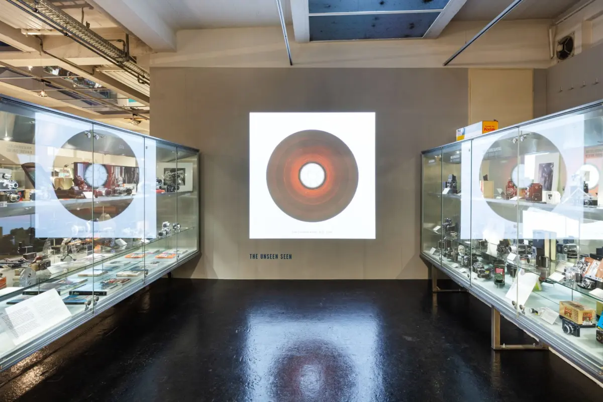 Gallery with glass display cases of vintage cameras on both sides; large illuminated poster showing a red circular lens, THE UNSEEN SEEN at bottom.
