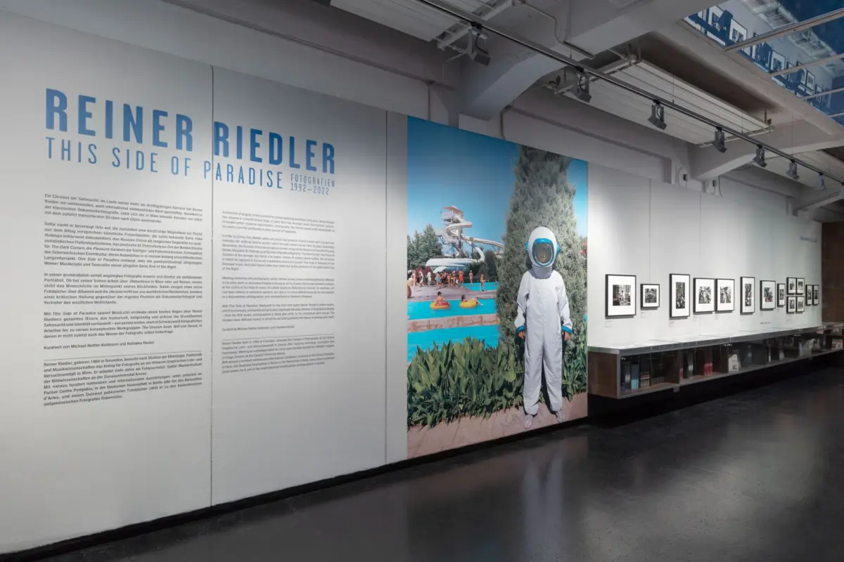 Indoor gallery wall for Reiner Riedler exhibition; large blue title and a person in a spacesuit beside a water-park photograph.