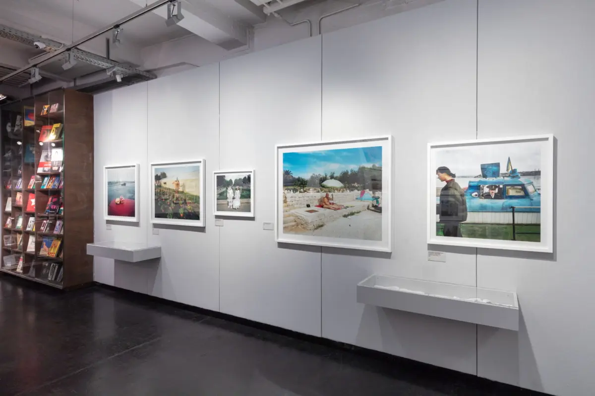 Art-gallery interior with framed photographs along a light-gray wall and a glass-fronted magazine shelf on the left.