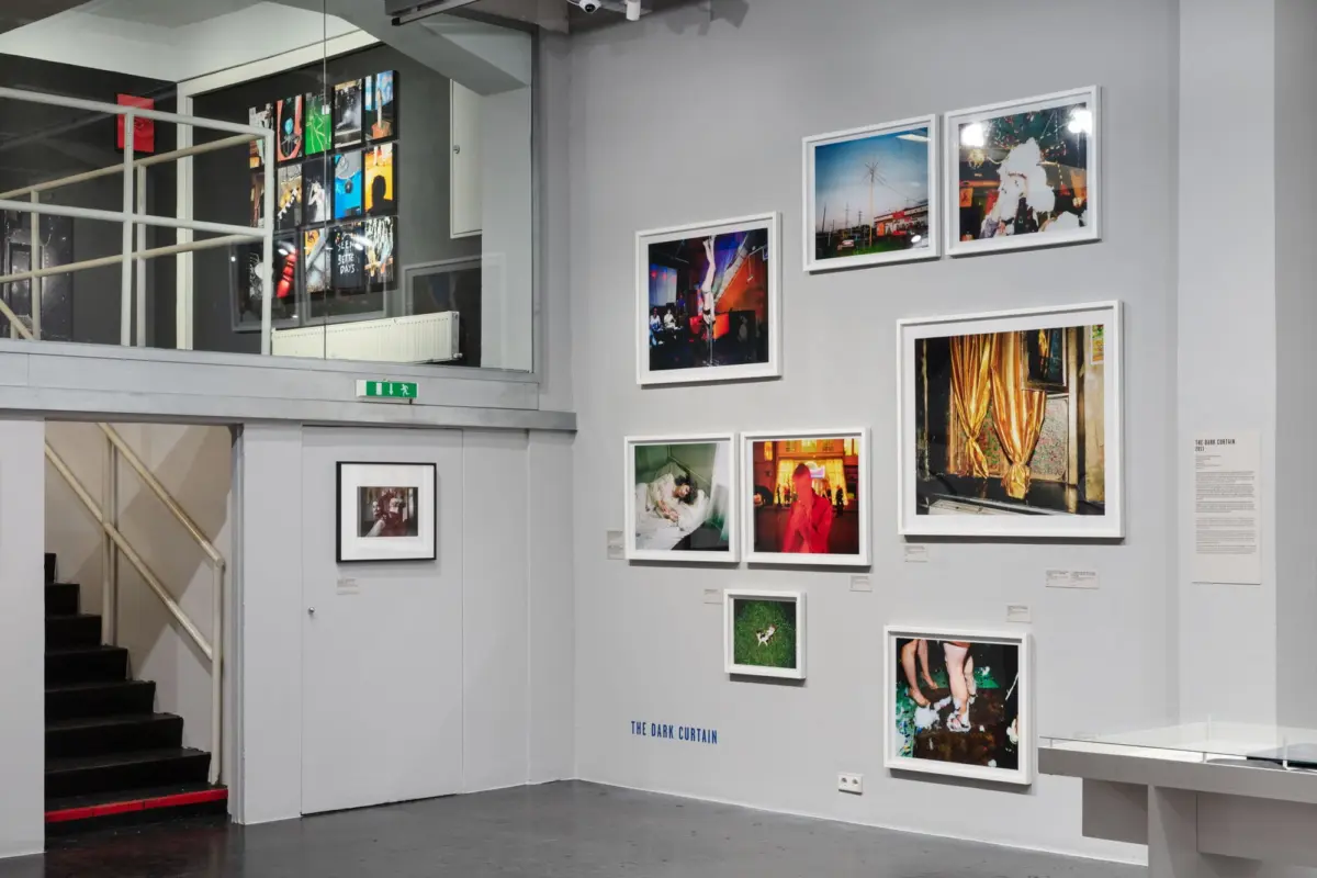 Interior of an art gallery featuring a wall of framed photographs arranged in a grid; staircase on the left.