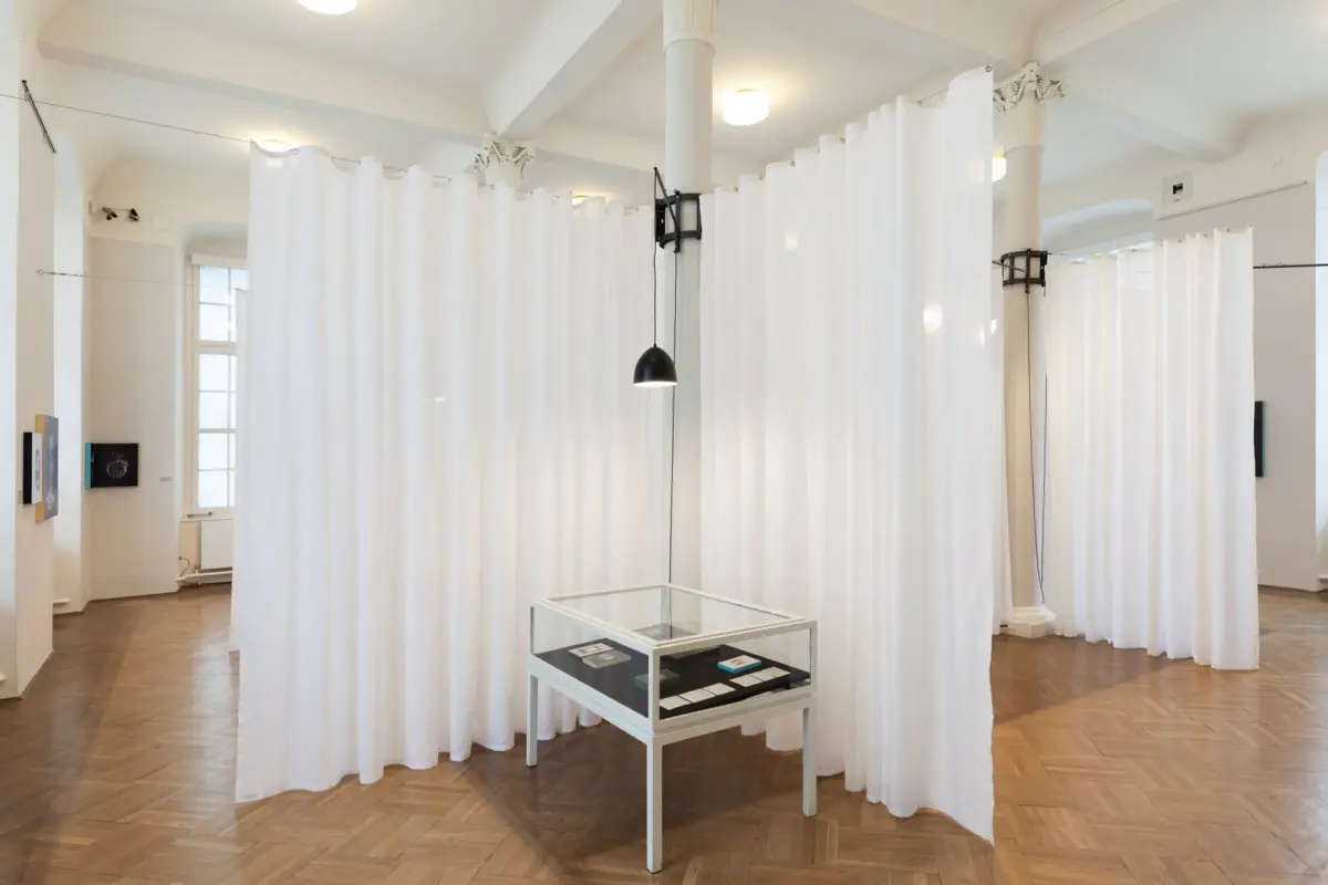 Art gallery interior featuring white curtains forming booths, ornate columns, a glass display case, and a black pendant lamp.