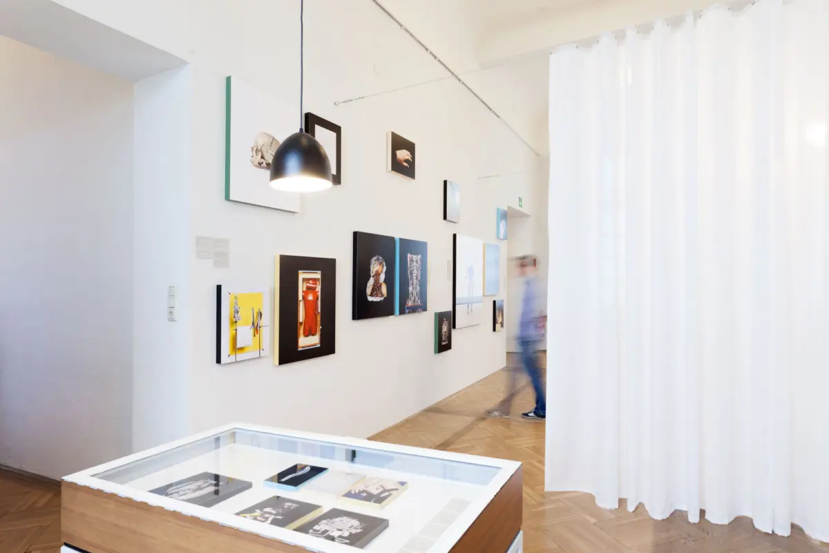 Art gallery interior with wall-mounted artworks, a glass-topped display case, and white curtains; a blurred visitor walking in the background.