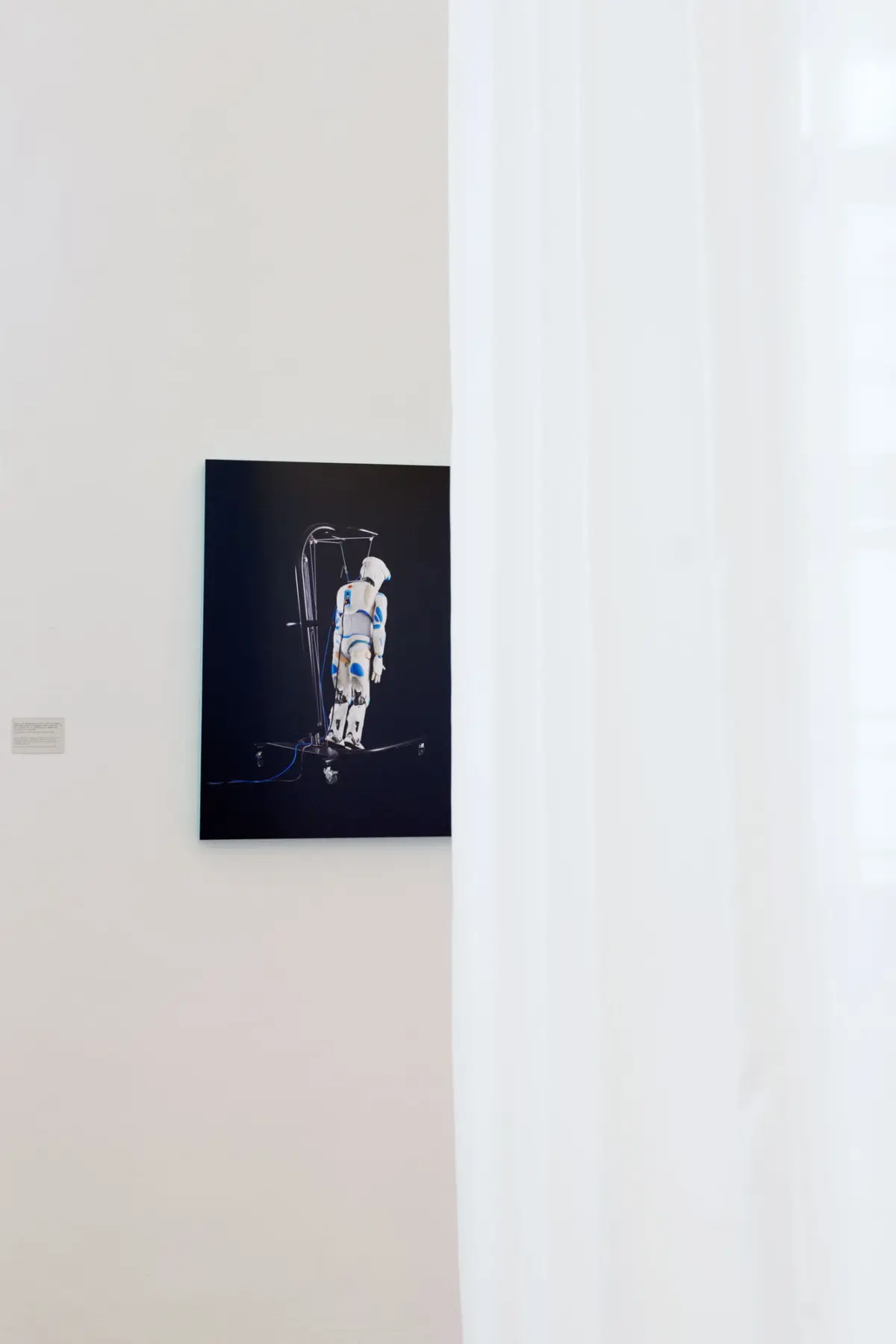 Poster of a humanoid robot on a wheeled stand displayed on a white gallery wall.
