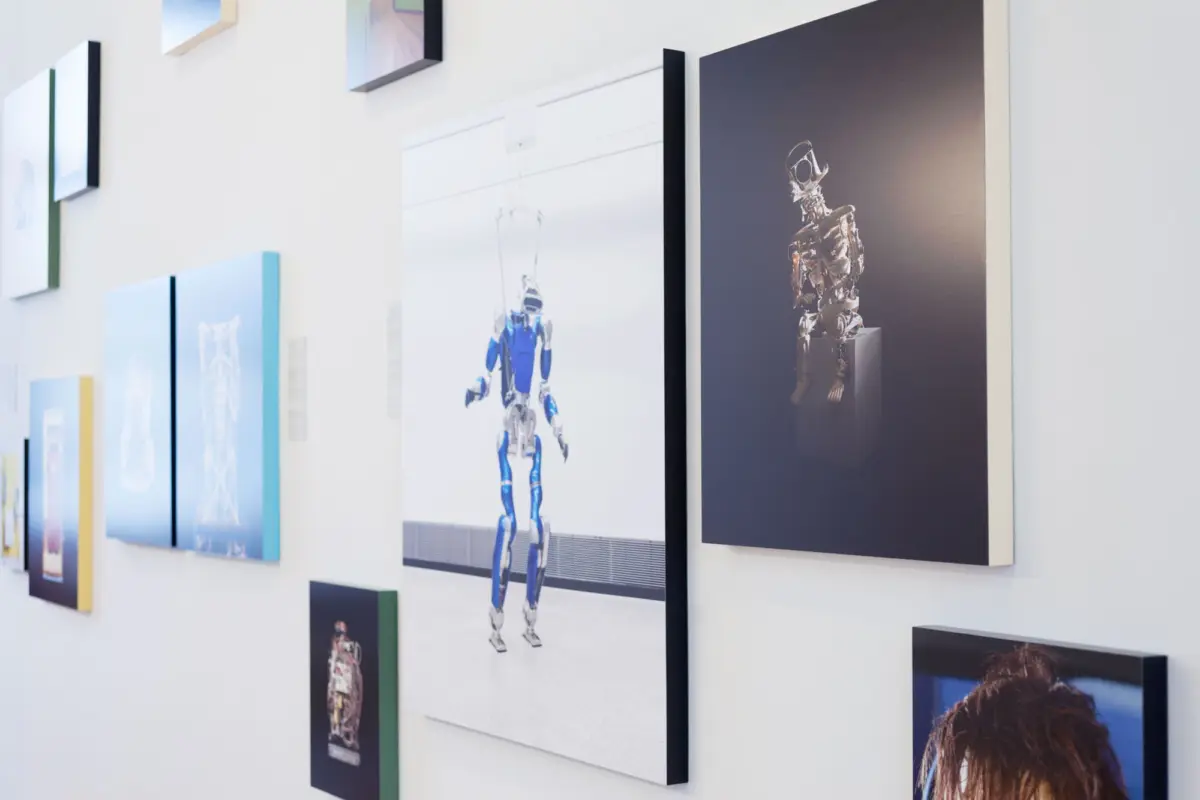 Blue humanoid robot standing on a large poster, displayed on a white gallery wall among multiple framed artworks.