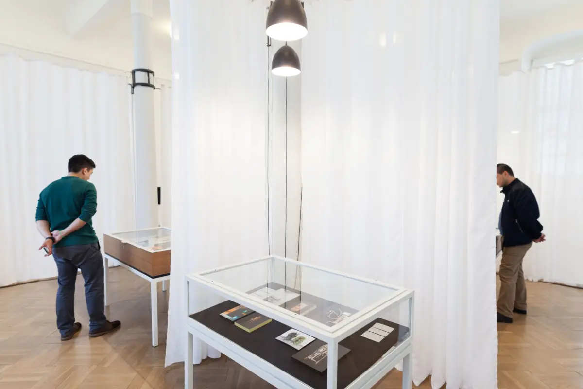 Two visitors in a bright gallery examine items in glass display cases amid white curtain partitions.