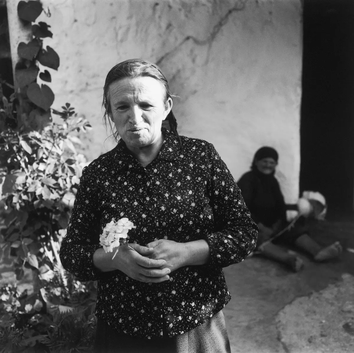 Older woman in a floral-patterned blouse holds a small bouquet outdoors beside a stone wall; two people sit in the background.