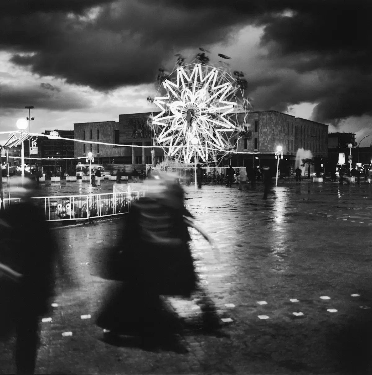 Illuminated starburst light sculpture in a rainy city square at night, with blurred pedestrians and wet reflections.