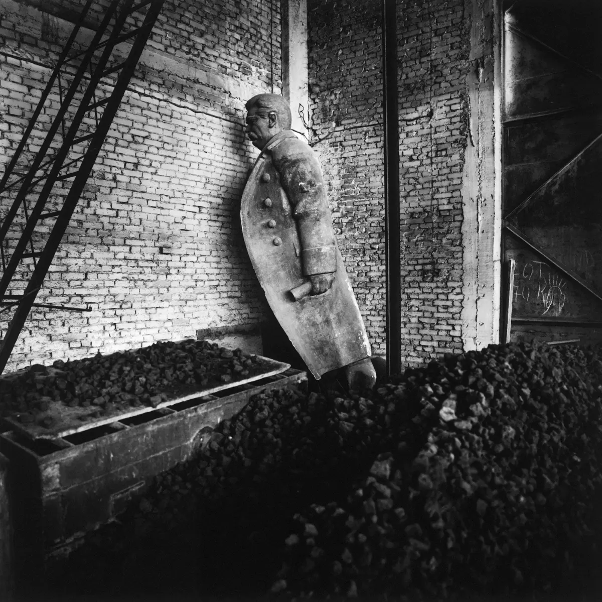 Man in a heavy overcoat leans against a brick wall in a coal yard, beside piles of coal and industrial equipment.