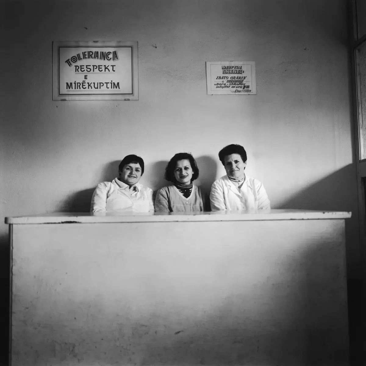 Three people in white coats sit behind a long desk against a textured wall in a room with wall-mounted signs.