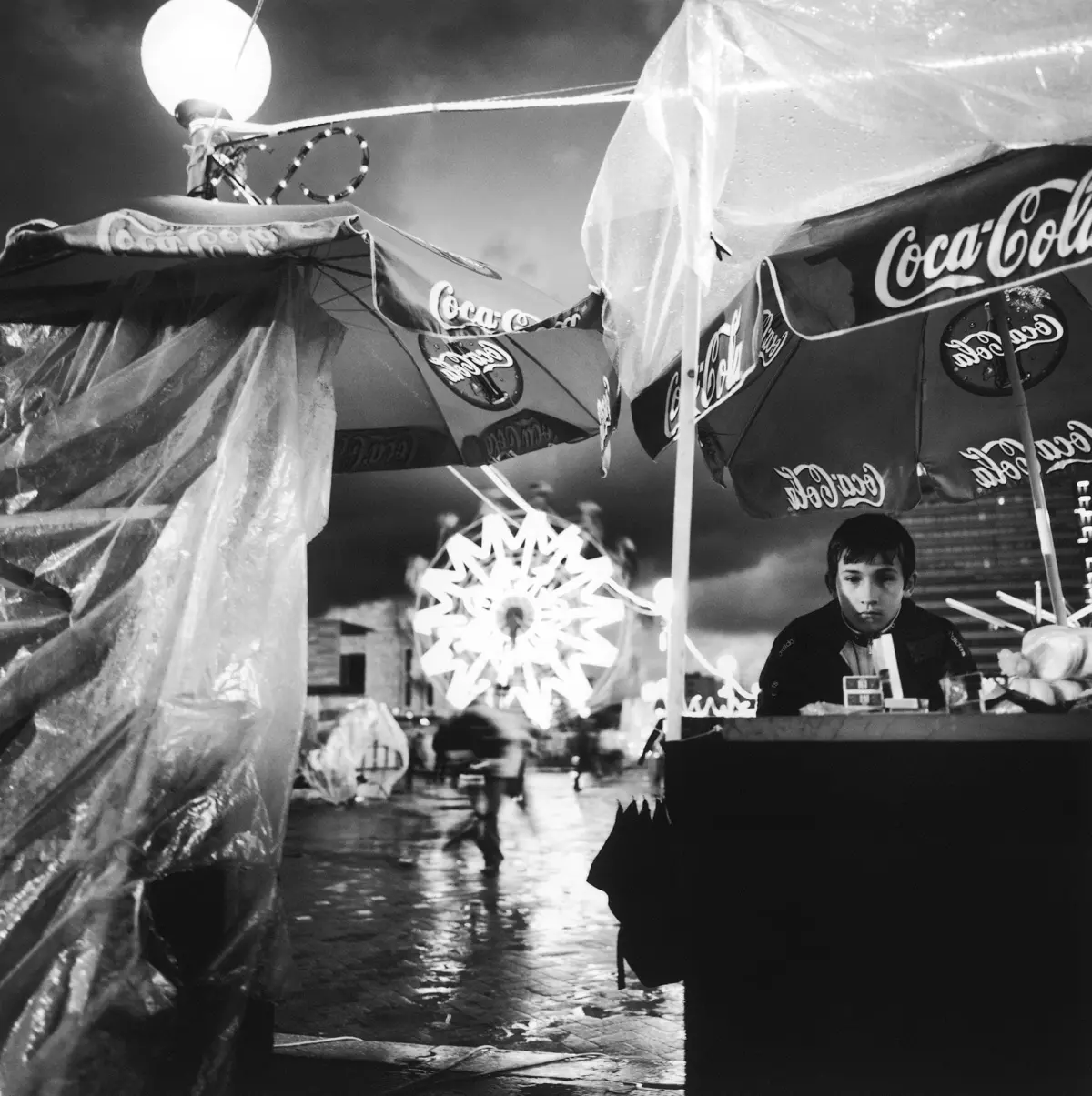 Vendor at a Coca-Cola umbrella stall on a rain-soaked street fair; bright star-shaped ride in the background.