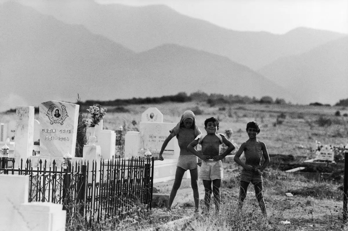 Three children stand with hands on hips beside tombstones in a rural cemetery, with distant mountains in the background.