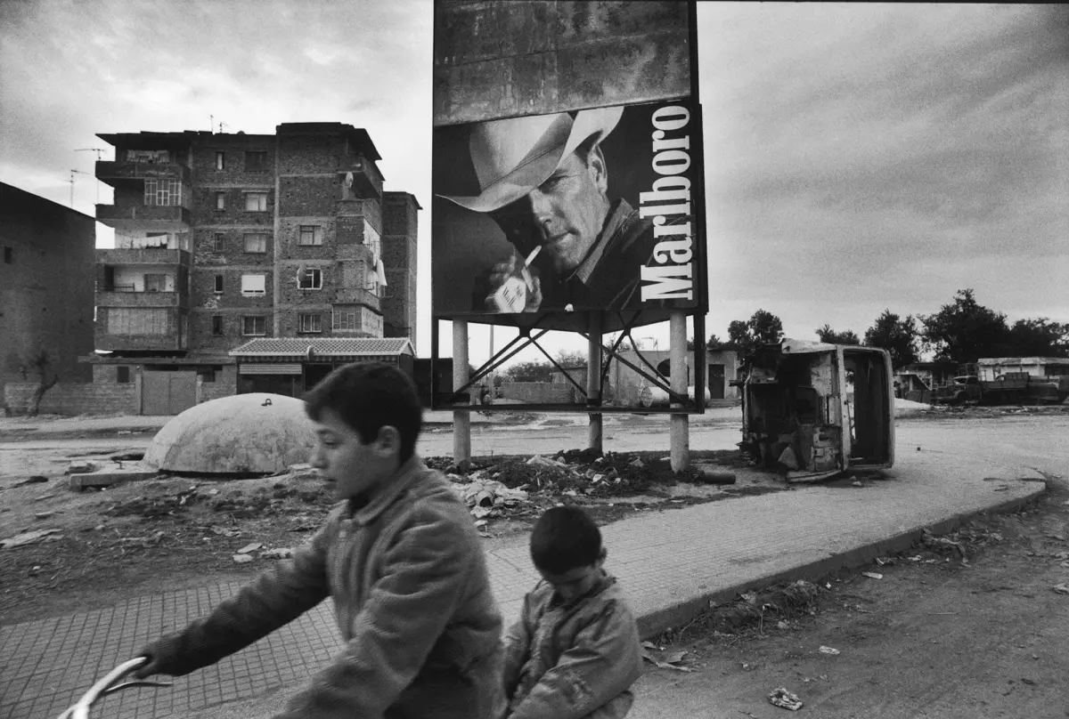 Two children ride a bicycle along a rubble-strewn street past a large Marlboro billboard in a damaged urban area.