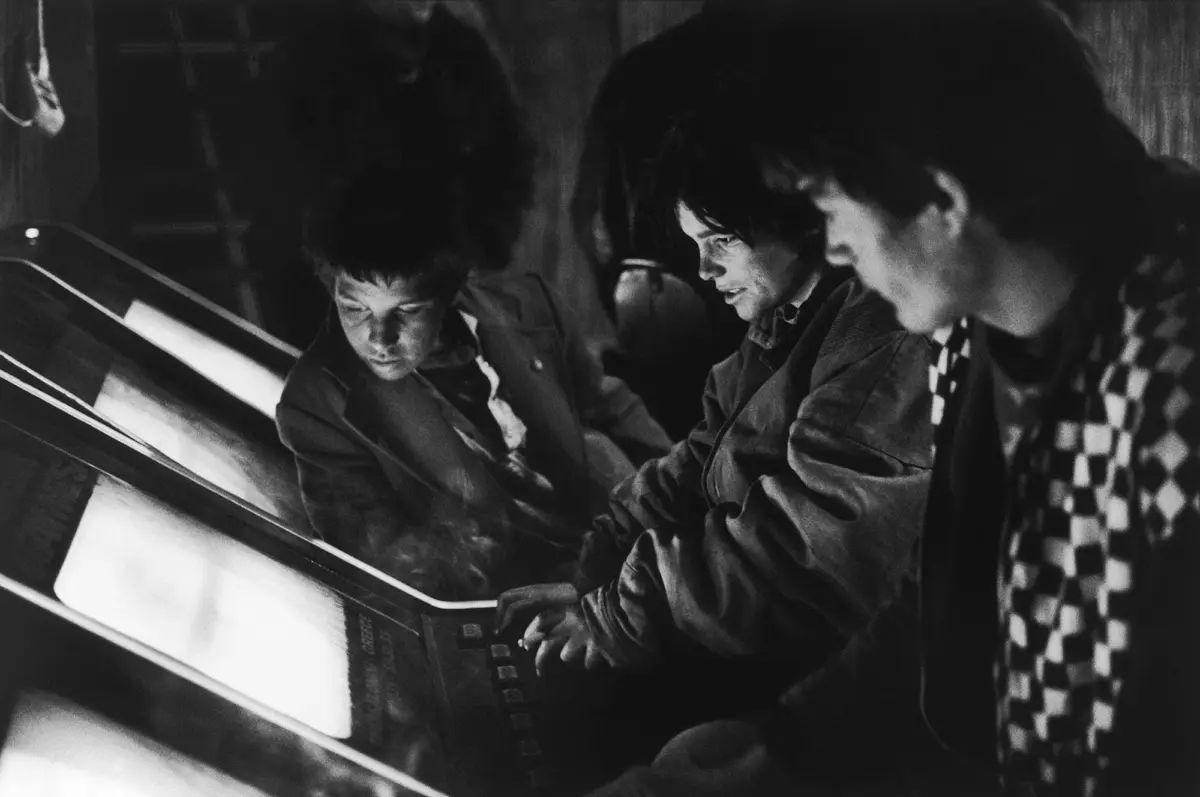 Three young people playing on upright arcade cabinets in a dimly lit arcade, focused on the screens.