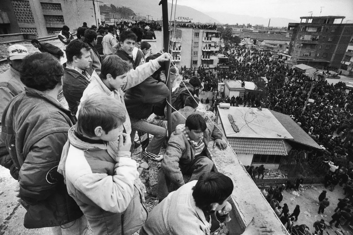 People on a rooftop watch a dense urban crowd filling streets below.