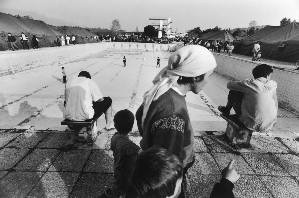 People sit and stand along the edge of an empty pool surrounded by tents in a refugee camp.